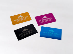 Fyra graverade metallkort i olika färger – gyllene, lila, svart och blå – med Norrgårdens logotyp.