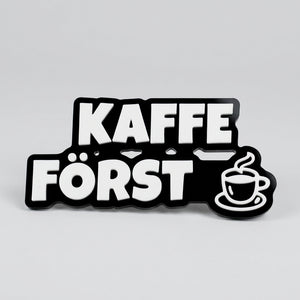Kylskåpsmagnet med texten ”Kaffe först” och en kaffekopp i vitt på svart bottenplatta