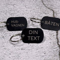 Tre nyckelringar i svart aluminium med graverad text "Husvagnen", "Båten" och "Din text", liggandes på betongliknande bakgrund.