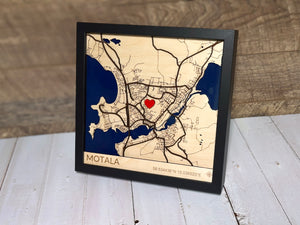 Handgjord karta i trä, laserskuren i flera lager med blåfärgat vatten, graverad stadsnamn och koordinater samt ett rött trä-hjärta som kan placeras valfritt.