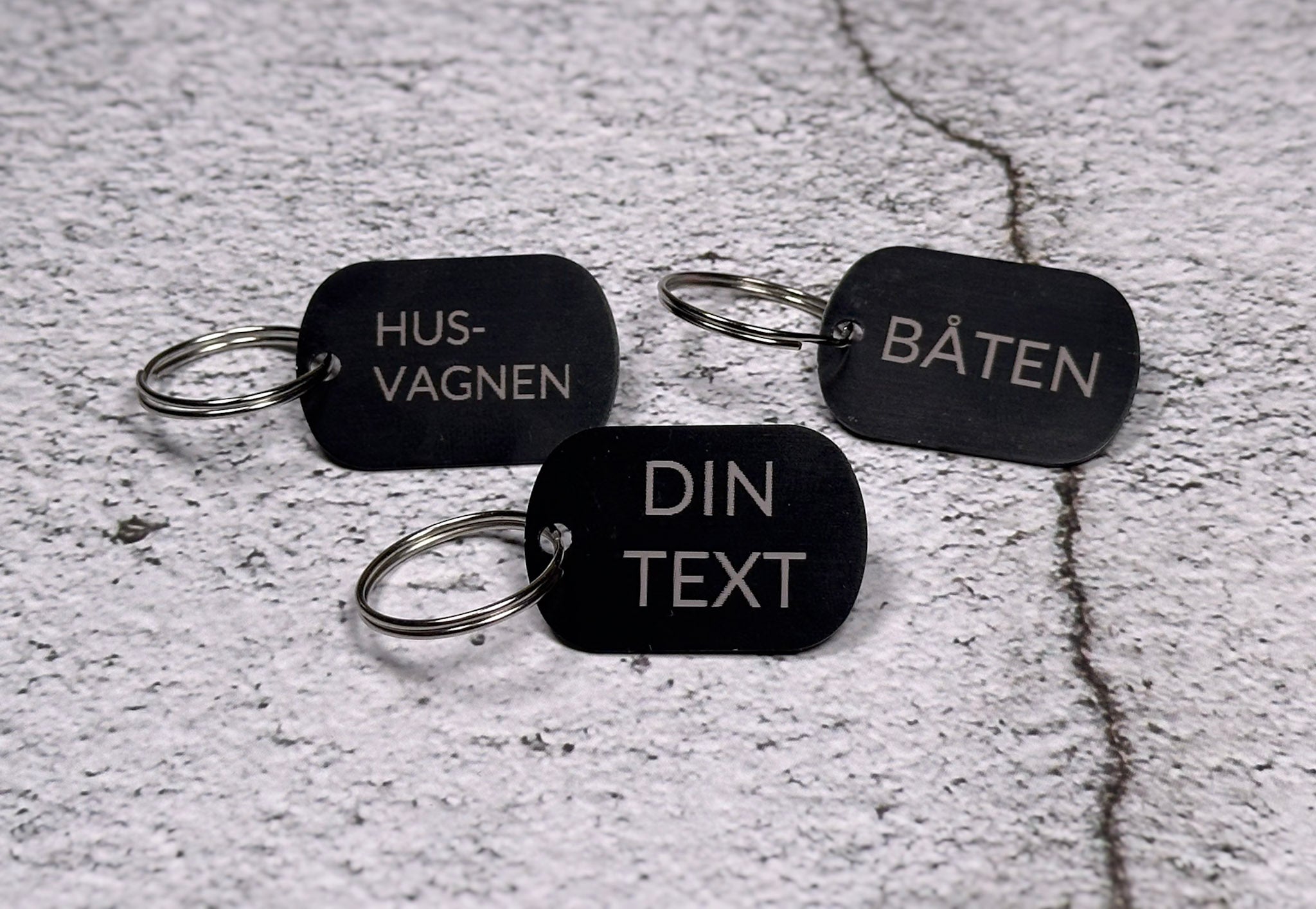 Tre nyckelringar i svart aluminium med graverad text "Husvagnen", "Båten" och "Din text", liggandes på betongliknande bakgrund.