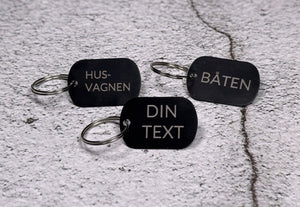 Tre nyckelringar i svart aluminium med graverad text "Husvagnen", "Båten" och "Din text", liggandes på betongliknande bakgrund.