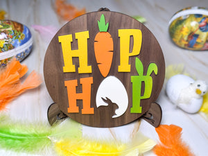 Rund träskylt med färgglad text 'HIP HOP', morot och kanin – placerad i påskmiljö